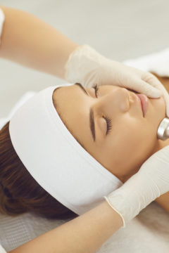 Cosmetologist making procedure of microdermabrasion of facial skin for young serene woman patient in neauty salon, top view. Cosmetology and professional skin care concept